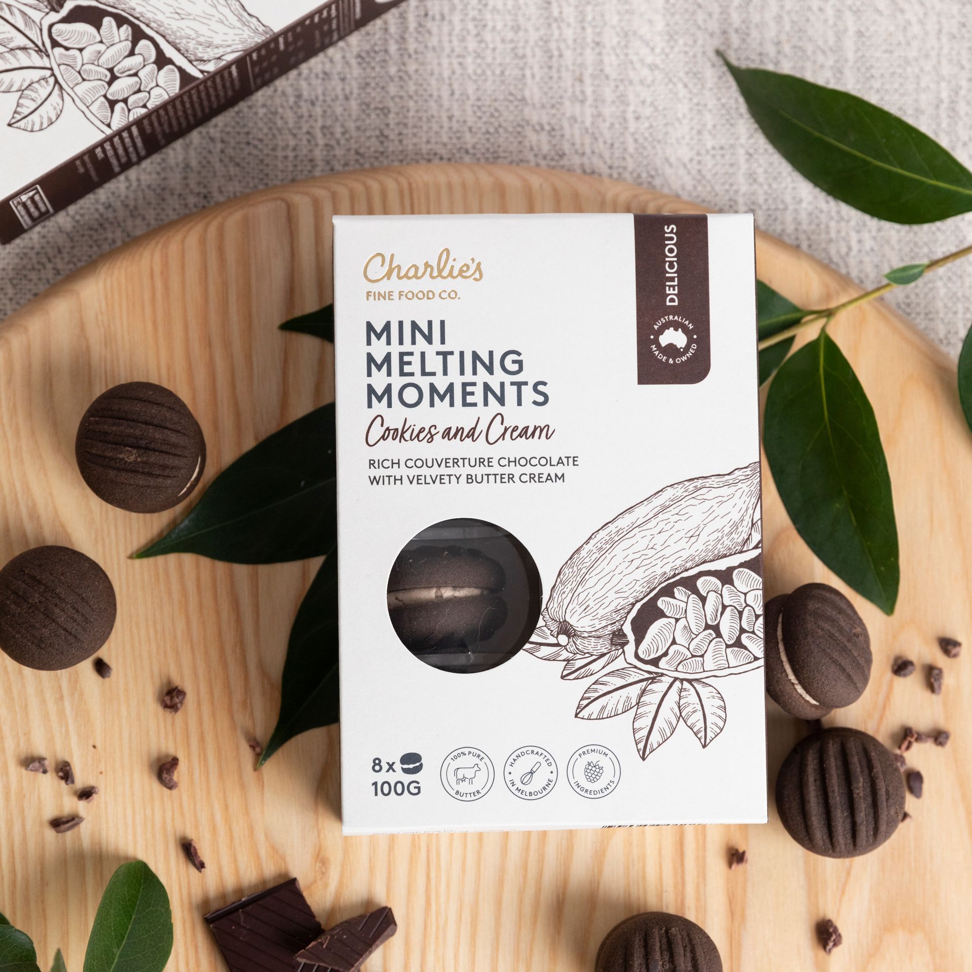 Box of Charlies Mini Melting Moments cookies on a wooden plate with scattered chocolate cookies and green leaves
