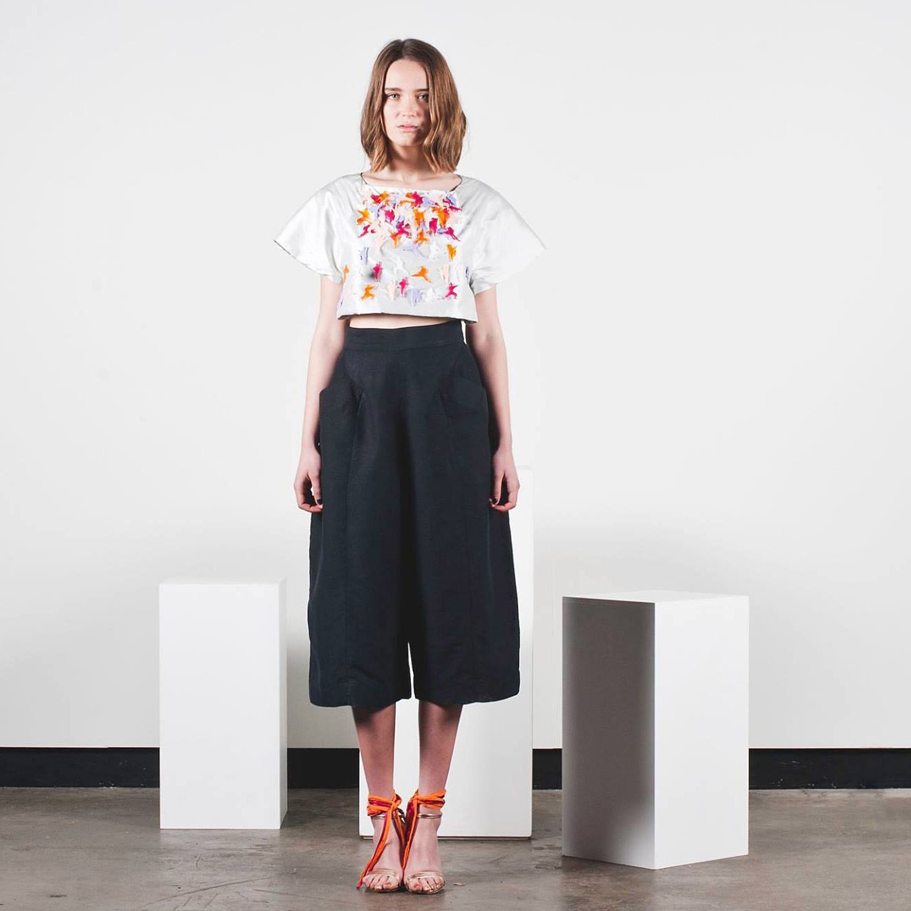 Woman standing in a minimalist studio wearing a white floral top, black wide-leg pants, and orange heels