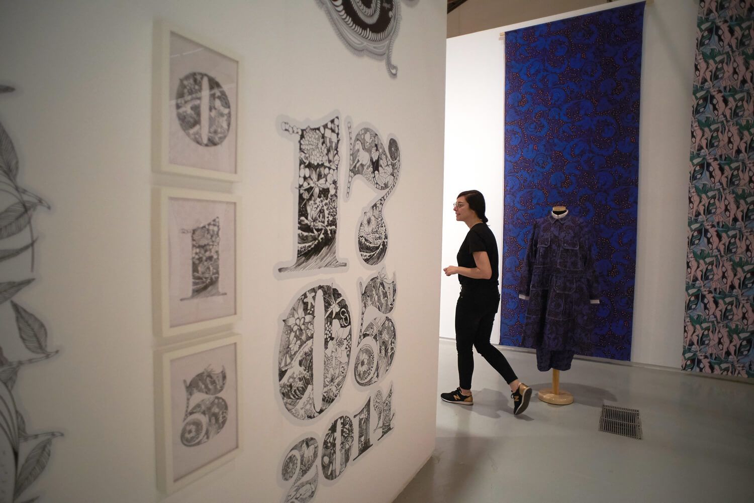 Art gallery with black-and-white prints on a white wall and a blue hanging artwork; a person stands nearby.