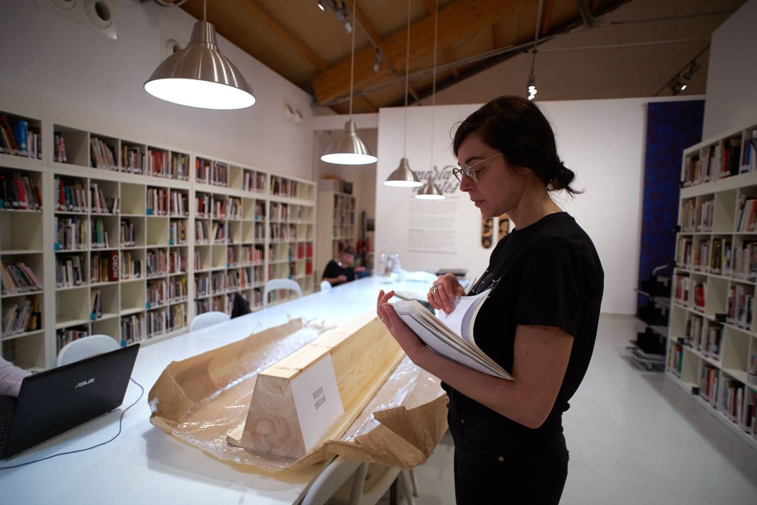 Woman taking notes beside a large wrapped object in a bright library or studio with bookshelves.
