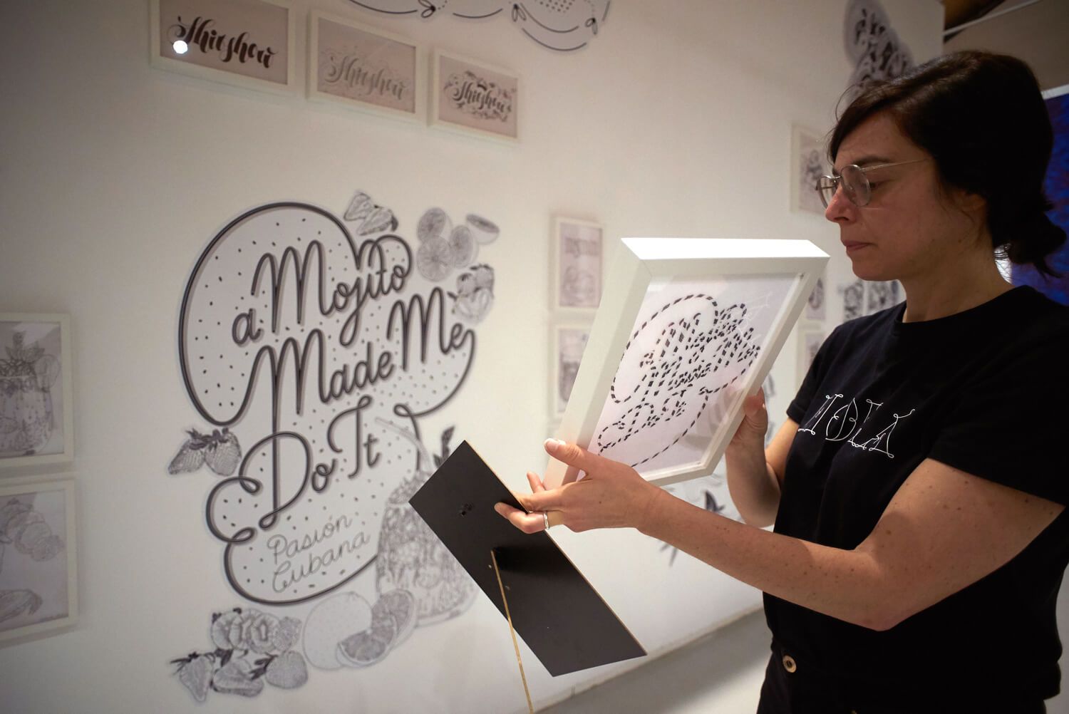 Woman in black shirt holding printouts beside framed calligraphy art in a bright studio
