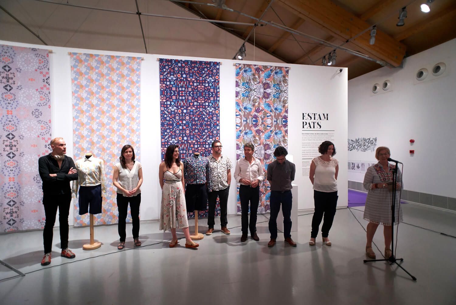 People lined up on a gallery stage before patterned textile panels and a white wall display.