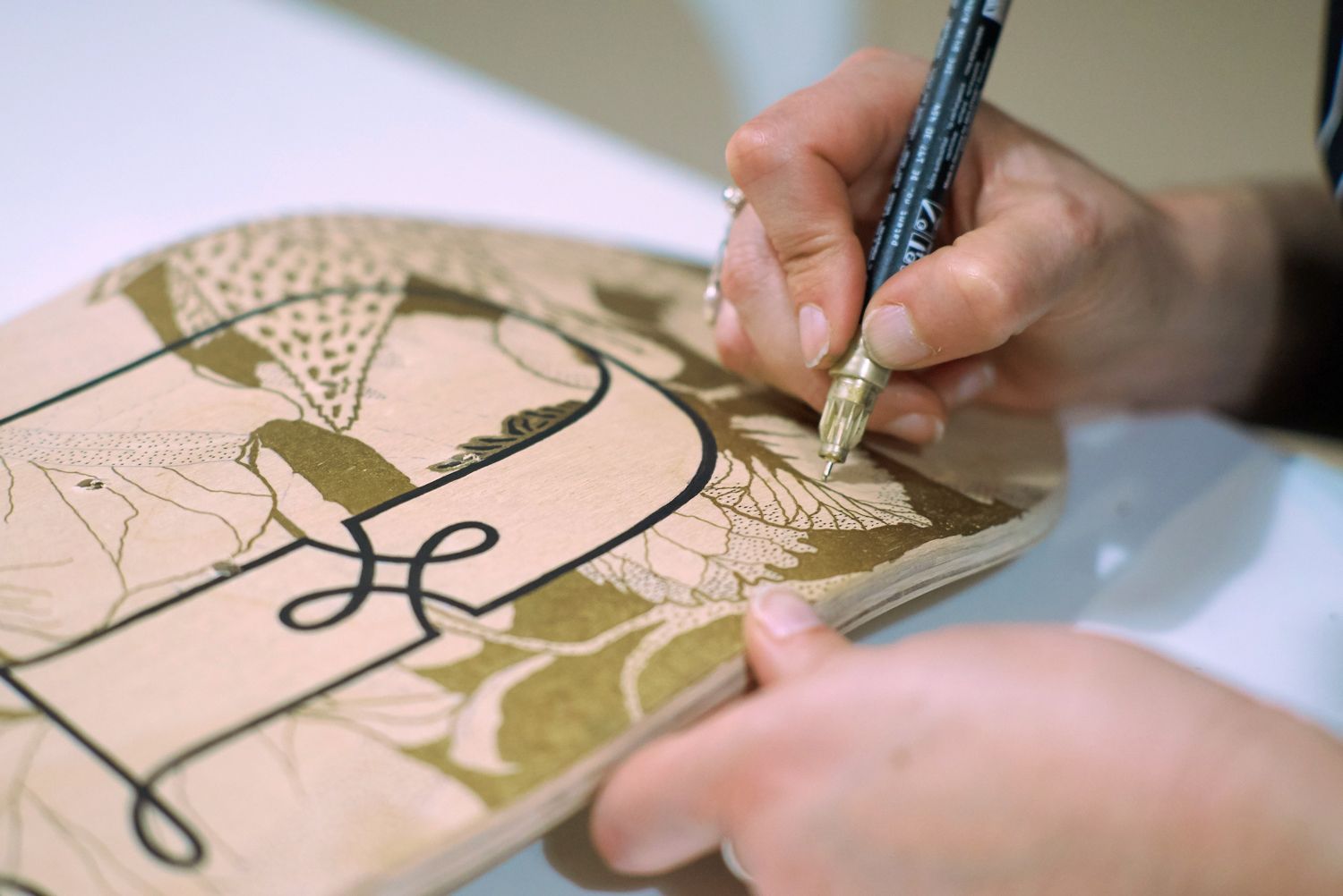 A person uses a fine-tipped black pen to outline a decorative floral design on a pale wooden surface.