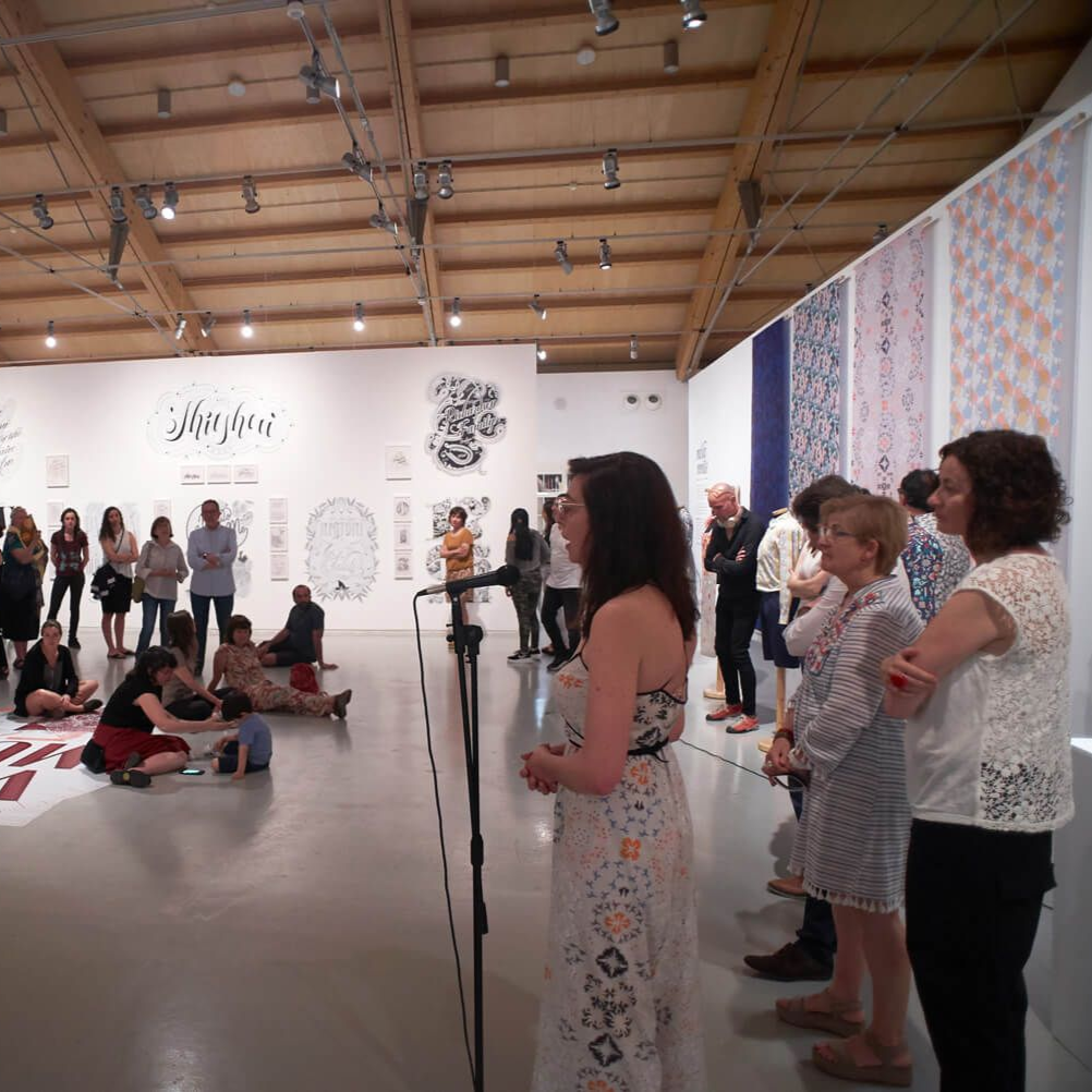 Visitors stand in a white gallery, watching a live performance under a wooden ceiling.