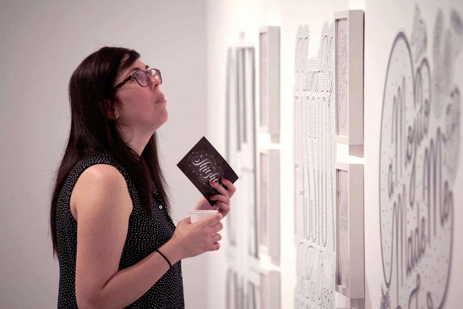 Woman in a gallery looking at framed artwork on a white wall, holding a brochure.
