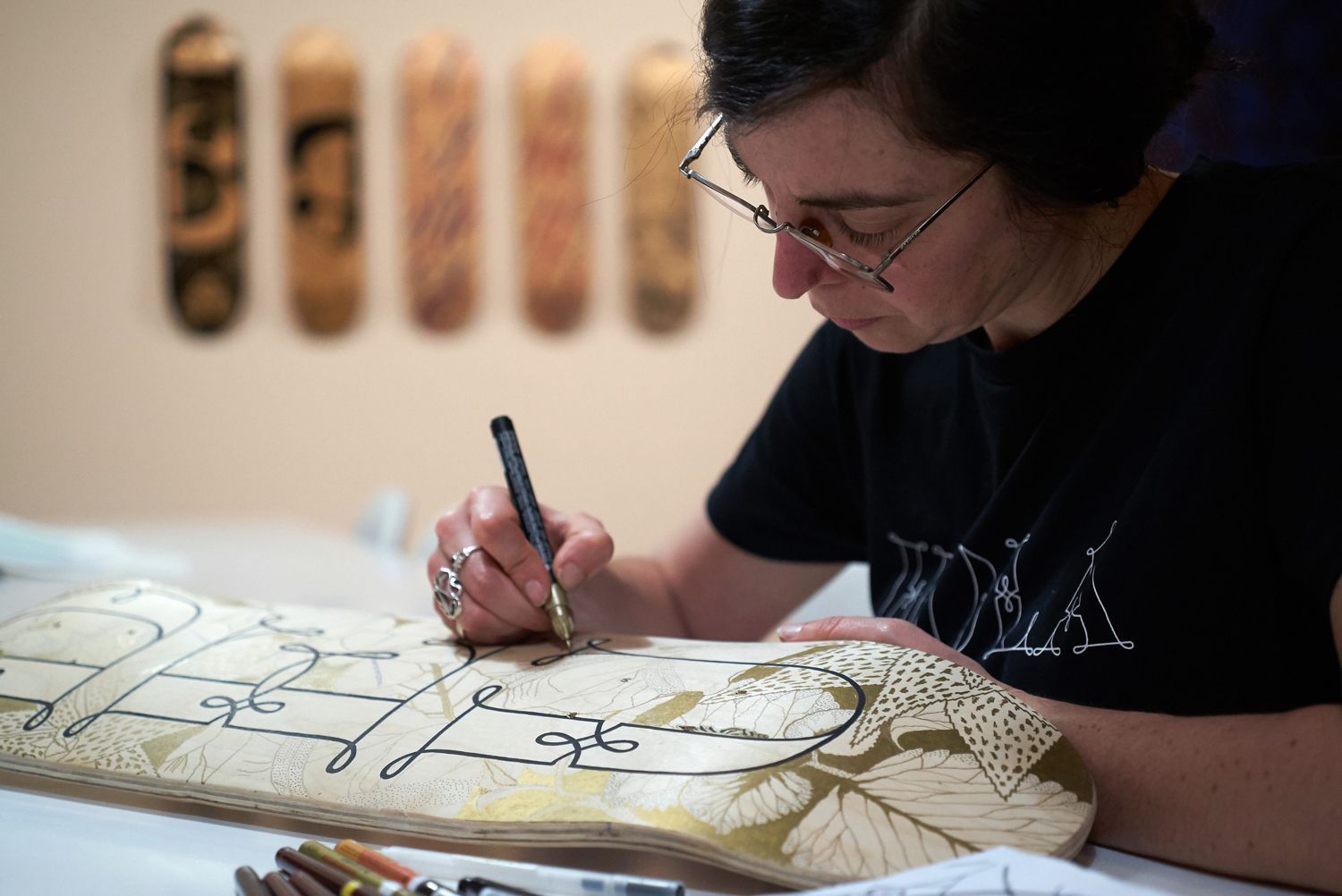 A person drawing a detailed design with a pen on a wooden skateboard deck, with finished boards hanging on the wall.