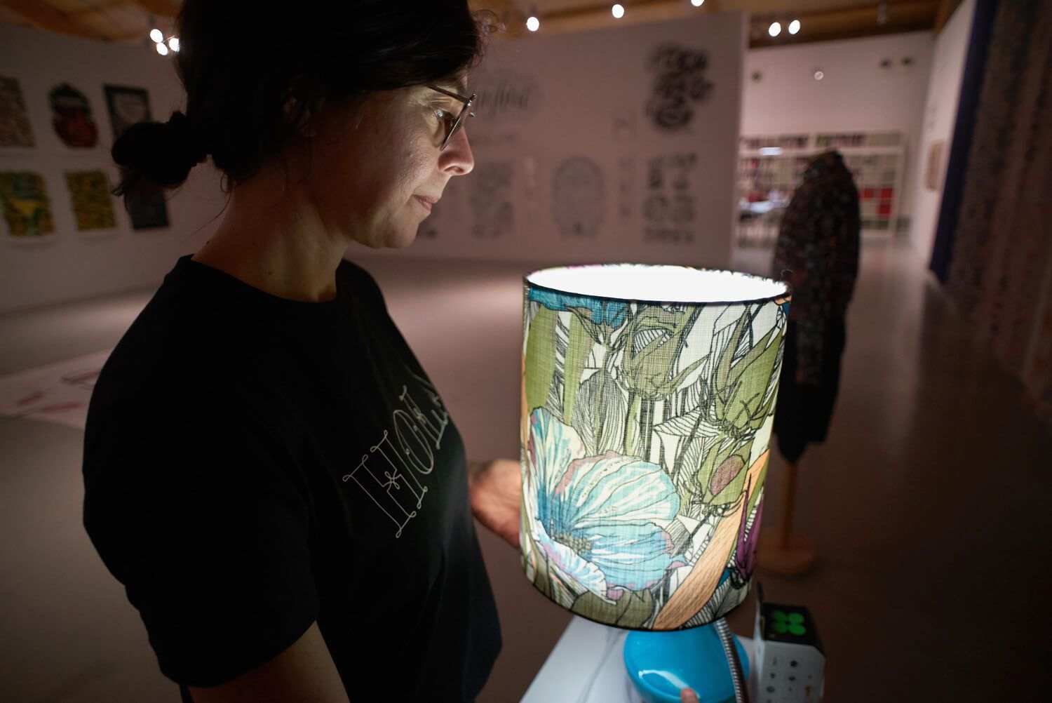 Person studying a glowing mosaic lamp in a dim room with posters on the wall