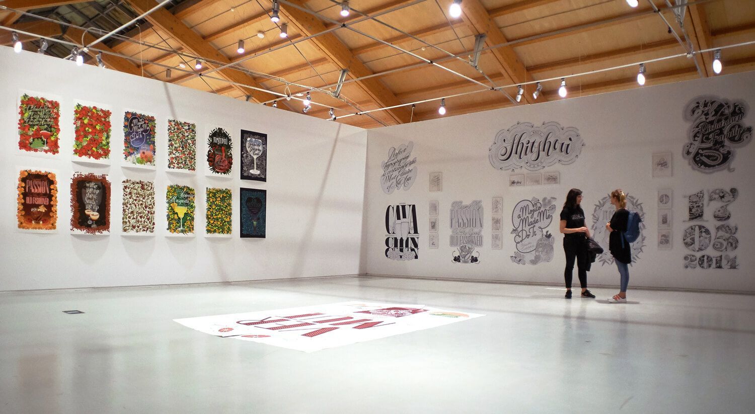 Art gallery with colorful wall prints, wood ceiling, and two people viewing large black-and-white artworks.