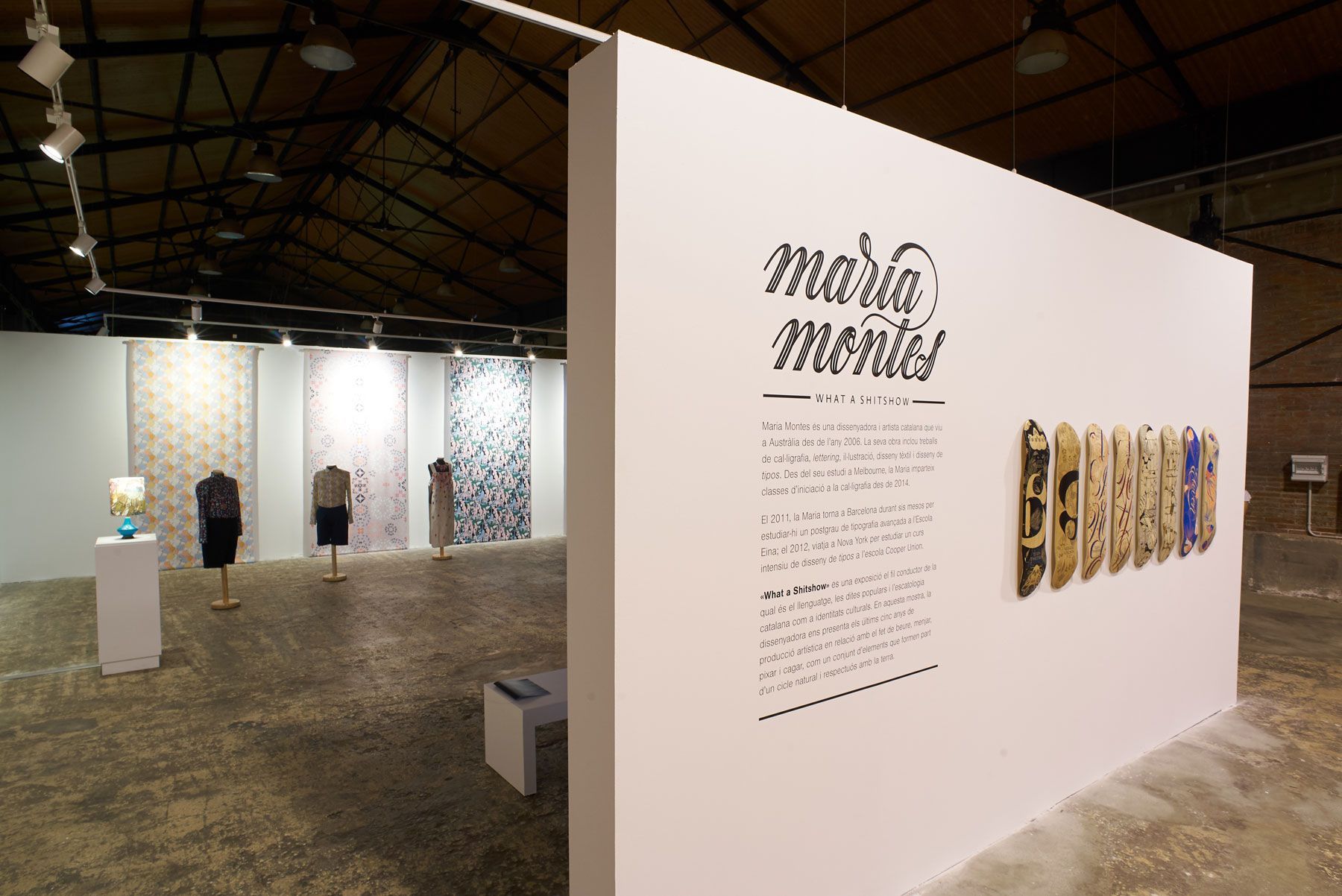 Art exhibition booth with white walls, text panels, and hanging artworks in a large indoor gallery.