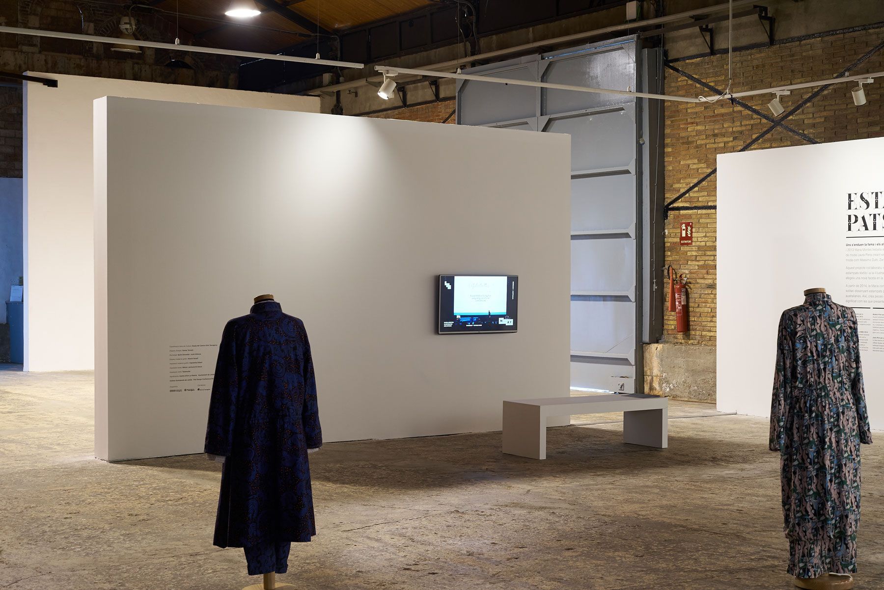 Minimal gallery exhibit with two hanging coats, a wall-mounted screen, and a bench in an industrial space