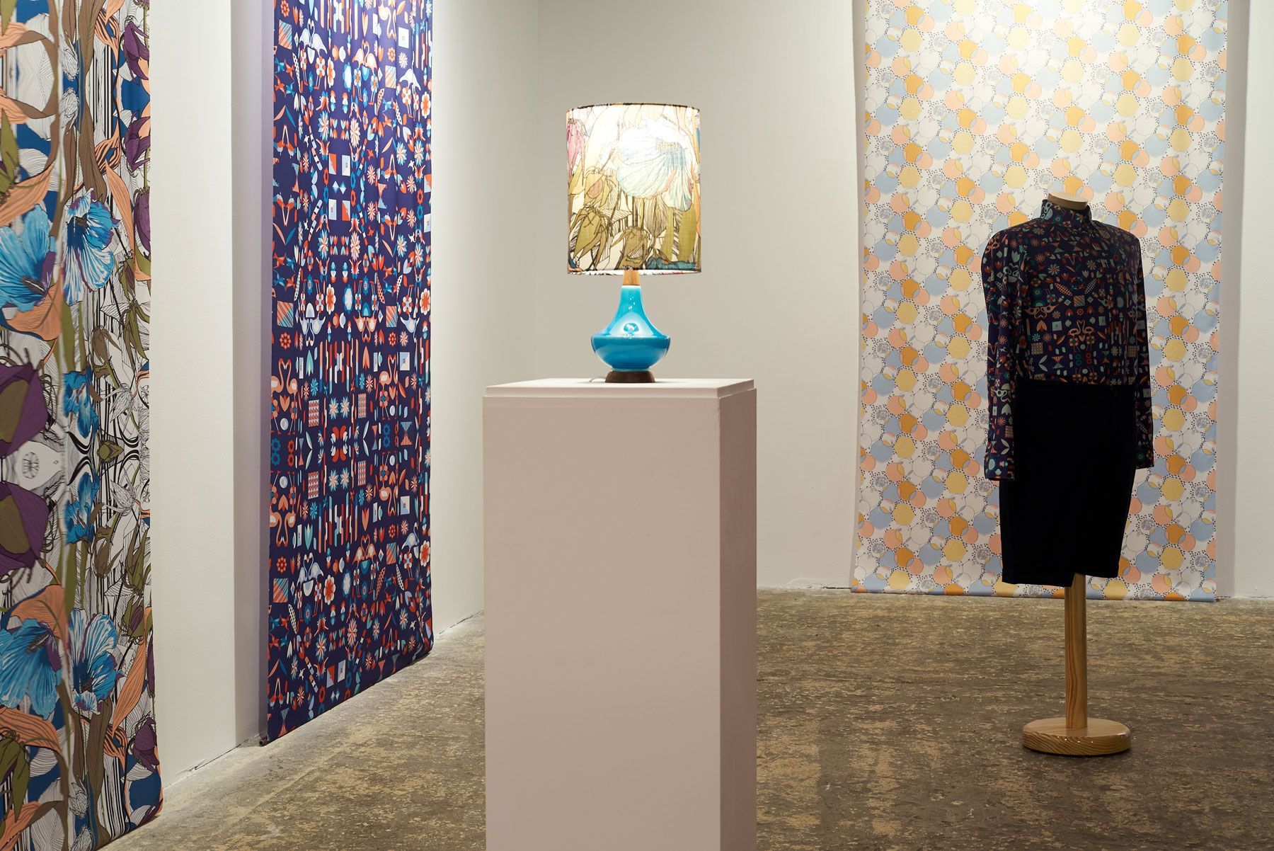 Art gallery with patterned textiles, a blue lamp on a pedestal, and a dark dress on a mannequin.