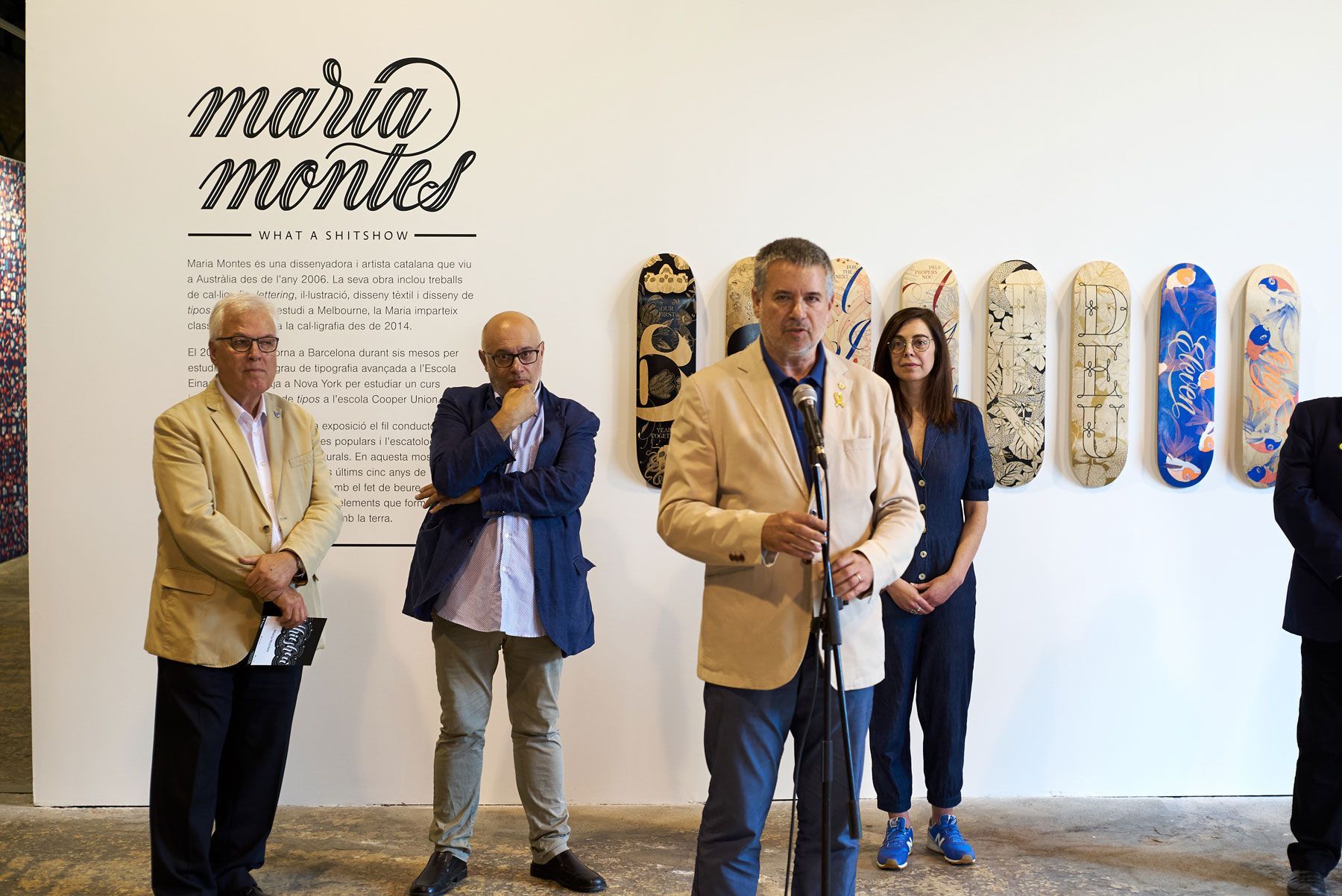 Four people stand in an art gallery beside skateboard decks mounted on a white wall.