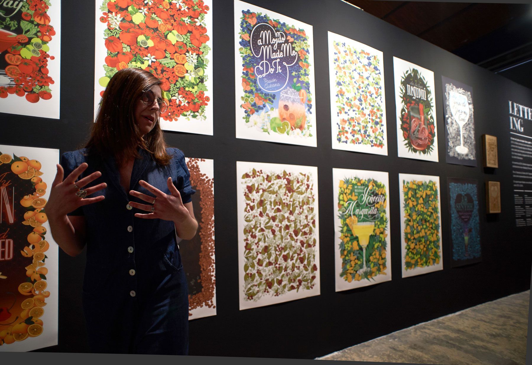 Woman speaking beside a wall of colorful framed art prints in a gallery exhibit