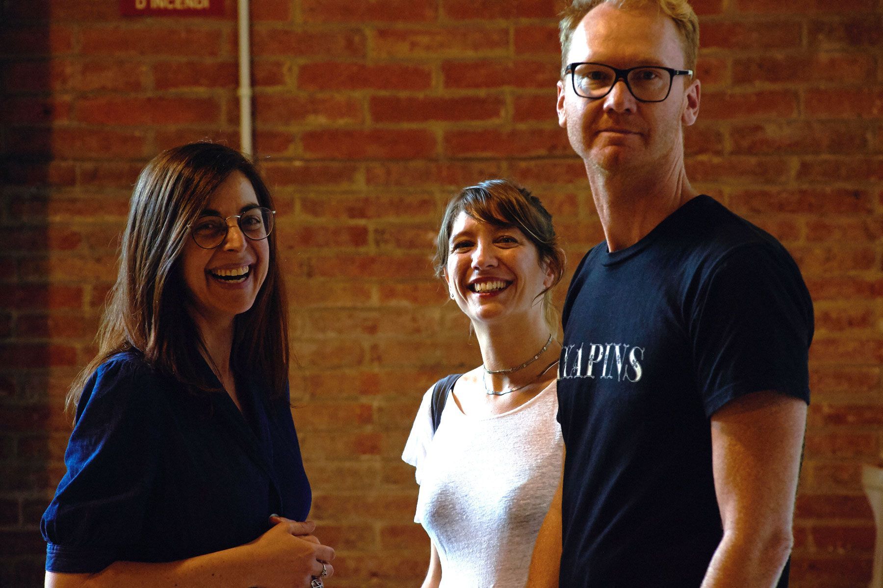 Three people smiling in front of a brick wall, with one wearing glasses and a black T-shirt.