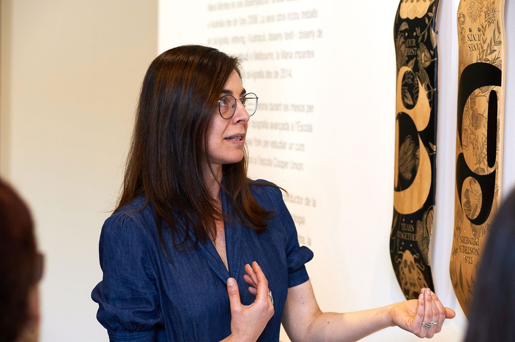 Woman in a blue shirt gestures beside wall-mounted carvings in a gallery.