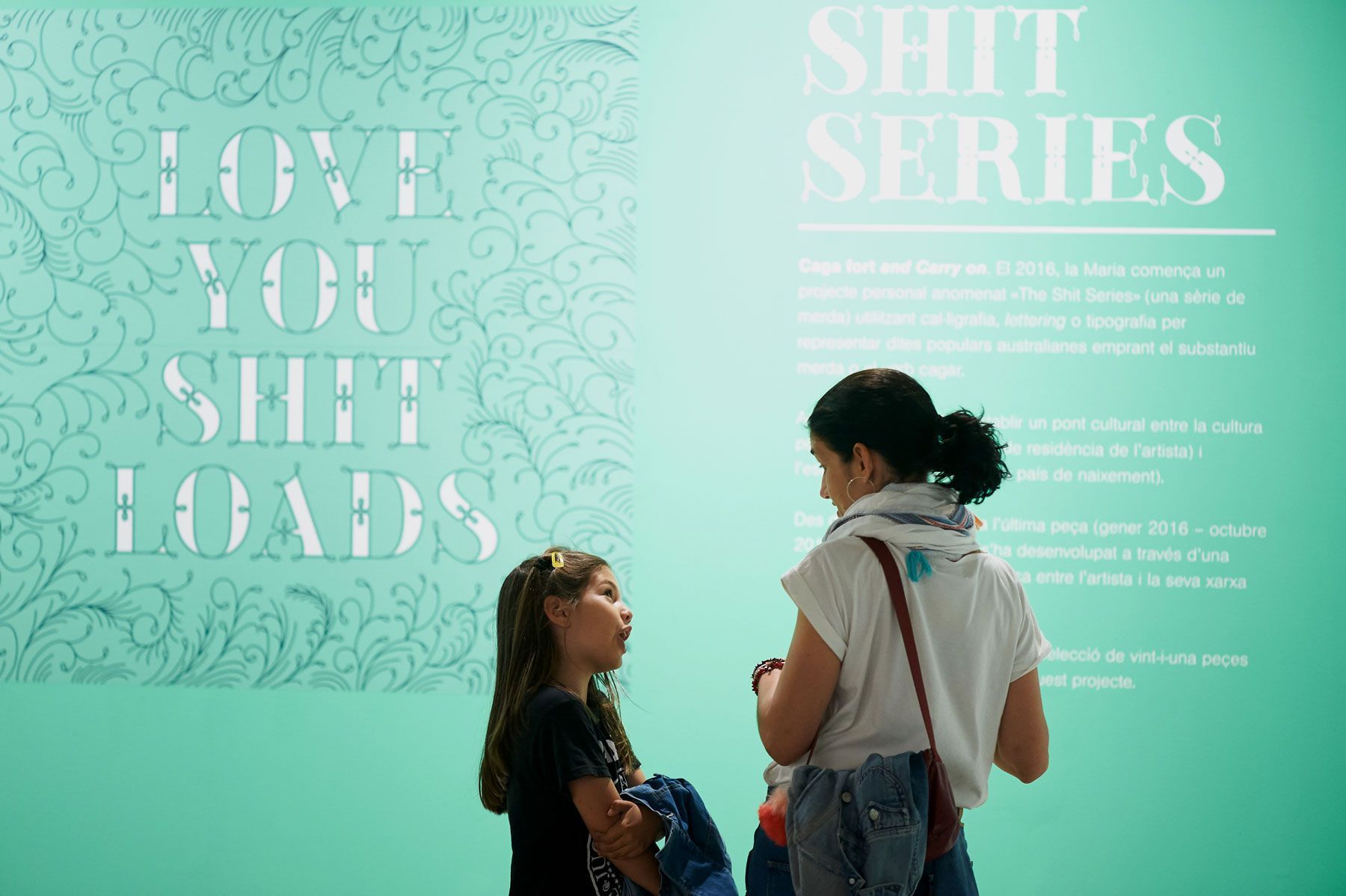 Two people stand before a turquoise wall with large white text reading “LOVE YOU SHIT LOADS” and “SHIT SERIES.”