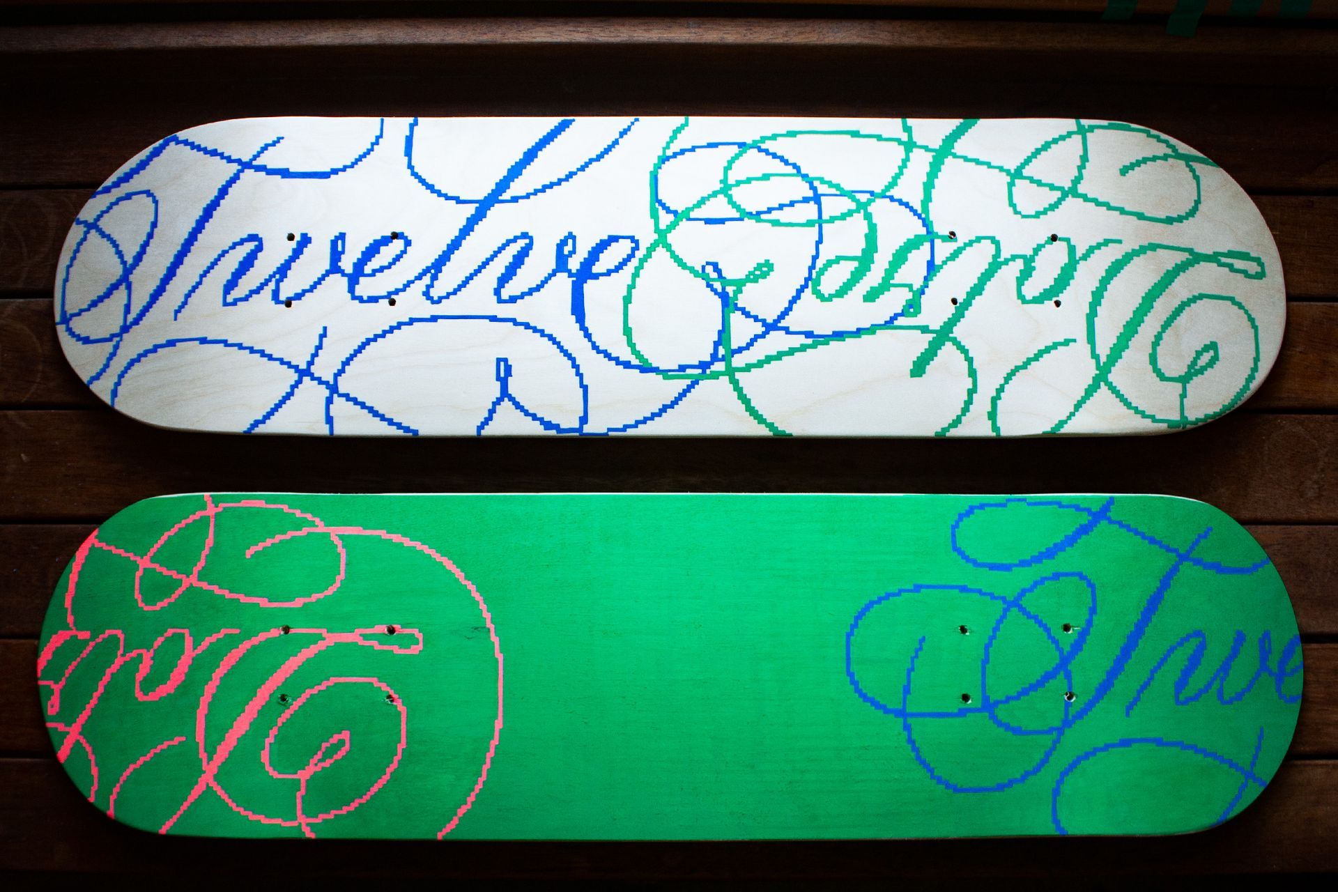 Two skateboards featuring cursive lettering, one with a white base and blue/green ink, the other green with blue/pink ink.