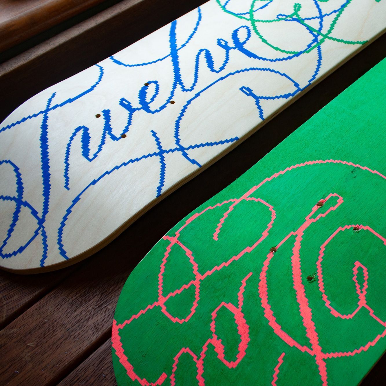 Two skateboard decks featuring stylized, cursive script: one white with blue text, the other green with pink text.
