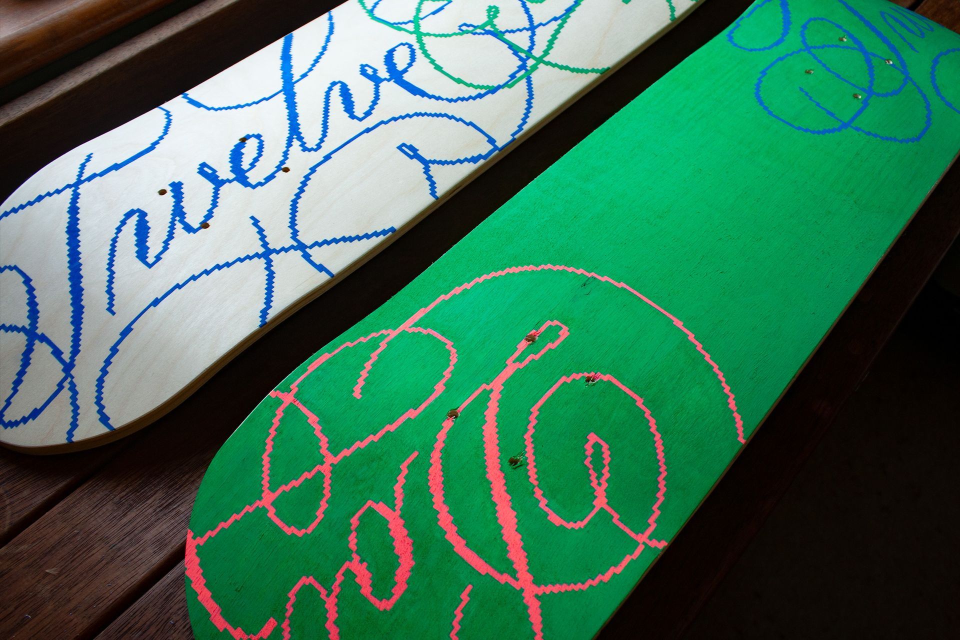 Two skateboards on a wooden surface: one white with blue script text, the other green with light pink script designs.