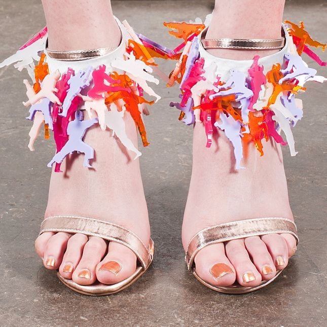 Feet in metallic sandals with colorful fringe ankle adornments on a gray floor