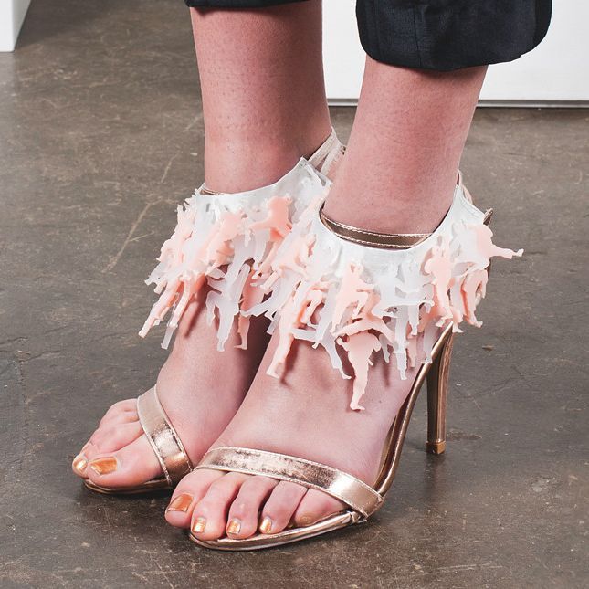 Feet in metallic gold sandals with pink feather ankle straps on a concrete floor.