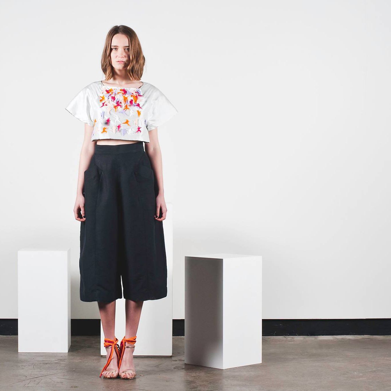 Model standing in a white studio, wearing a floral top, dark wide-leg pants, and red heels.
