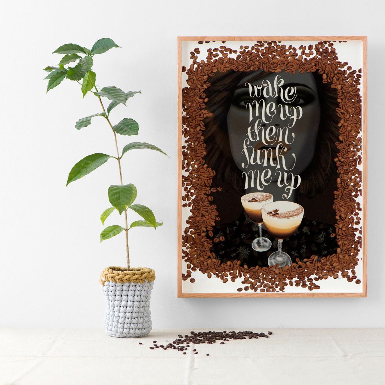 Framed coffee art print with brown beans, latte cups, and cursive text beside a potted vine on a white wall