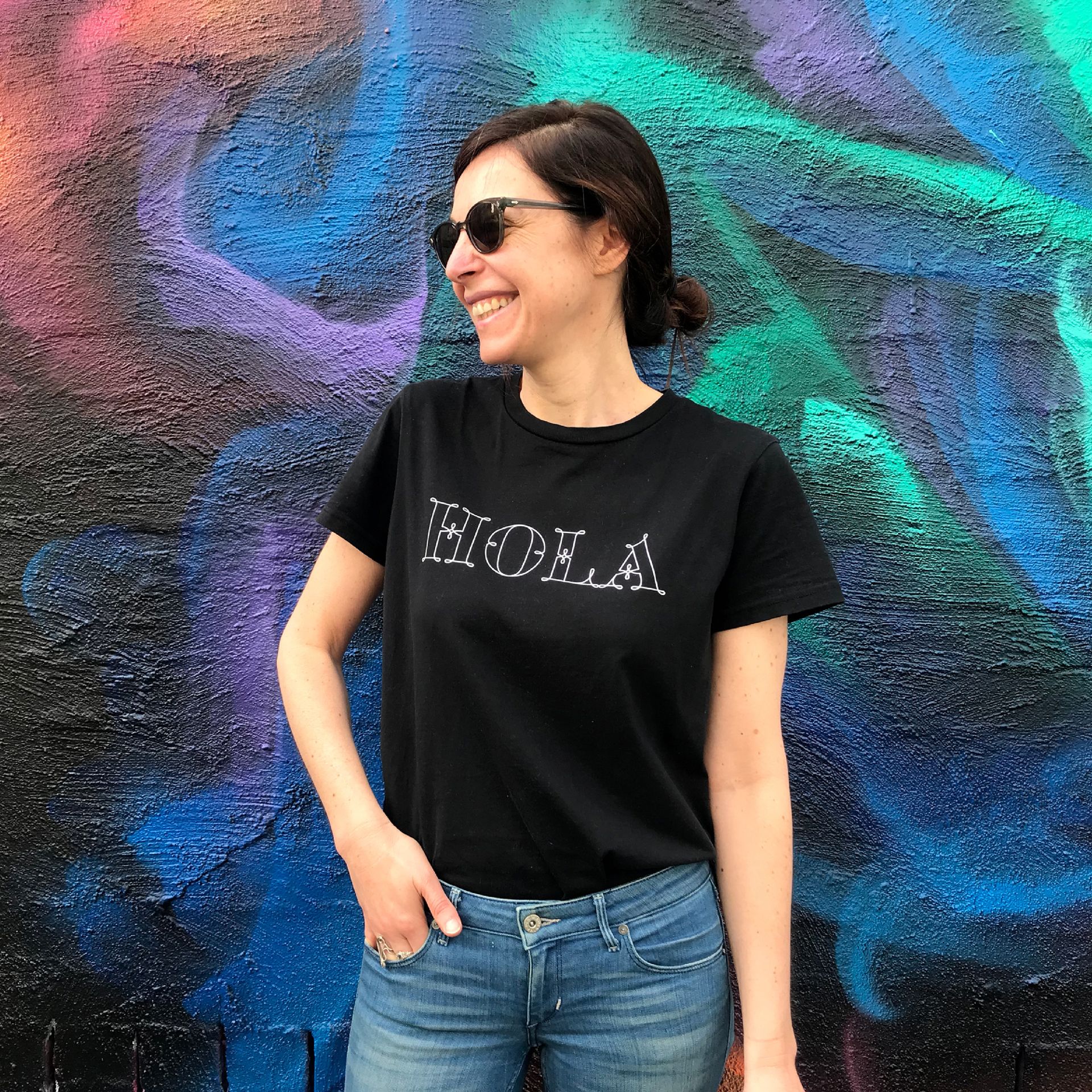 Green Fairy Font. Maria Montes in sunglasses and black T-shirt standing before a colorful graffiti wall and smiling.