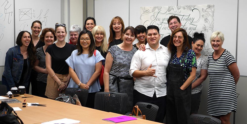 A large group of people stand together and smile in an office setting, with a whiteboard in the background.