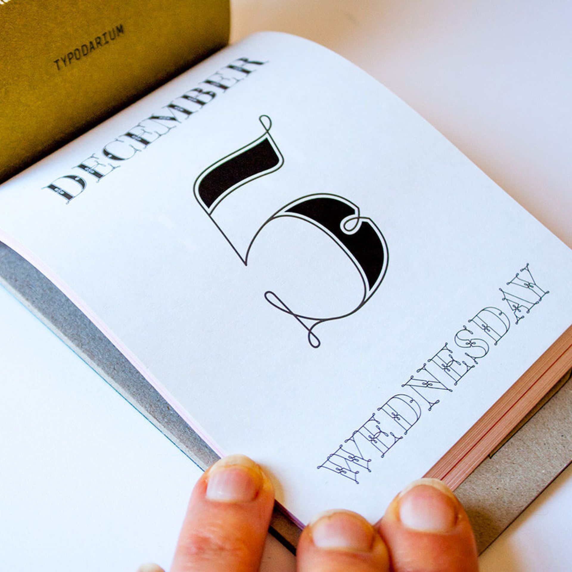 Hand holding open notebook with stylized black number 5 and the words “DECEMBER” and “WEDNESDAY” on the page