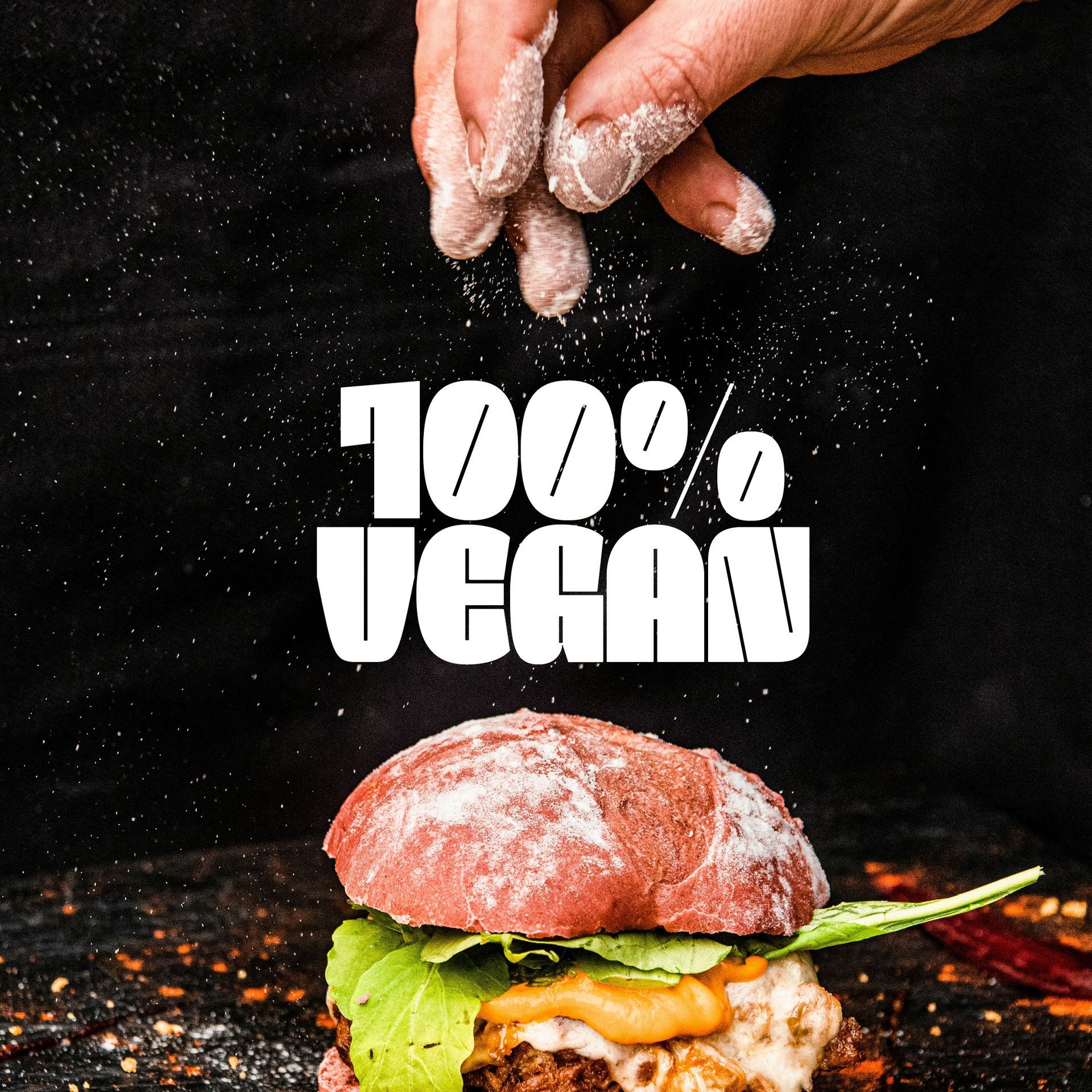 Hand sprinkling flour over a burger with “100% VEGAN” text on a black background