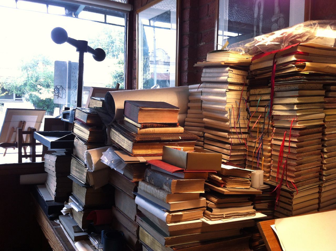 Stacks of books piled in a bookstore or library by a window with a lamp and shelves.