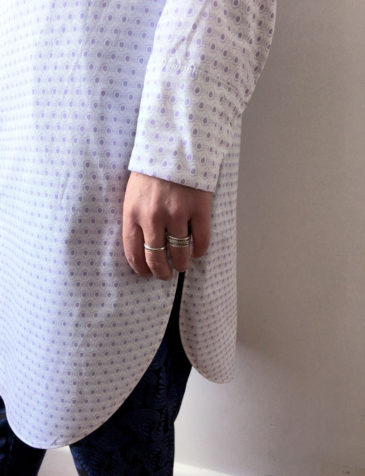 Person in a white patterned shirt with rings, standing against a light wall.