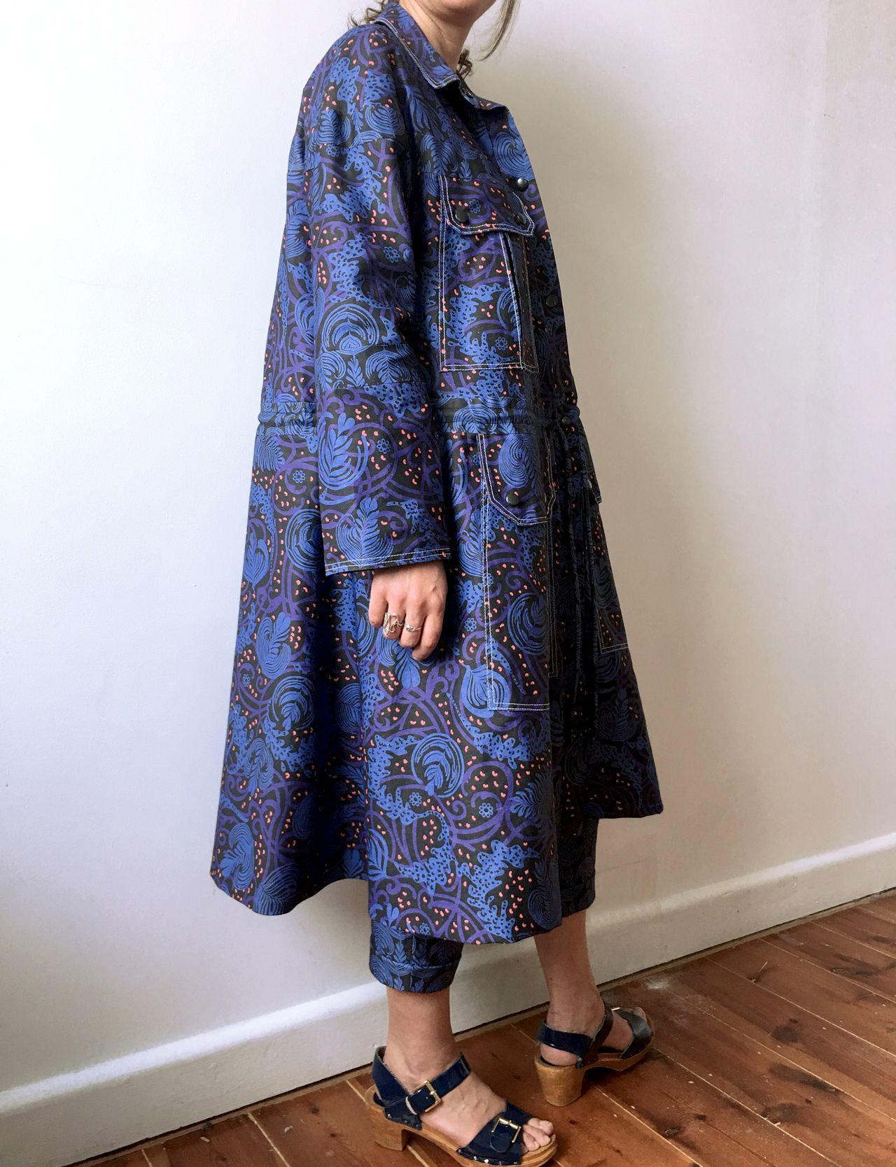 Person wearing a blue patterned coat and sandals standing by a white wall and wooden floor.