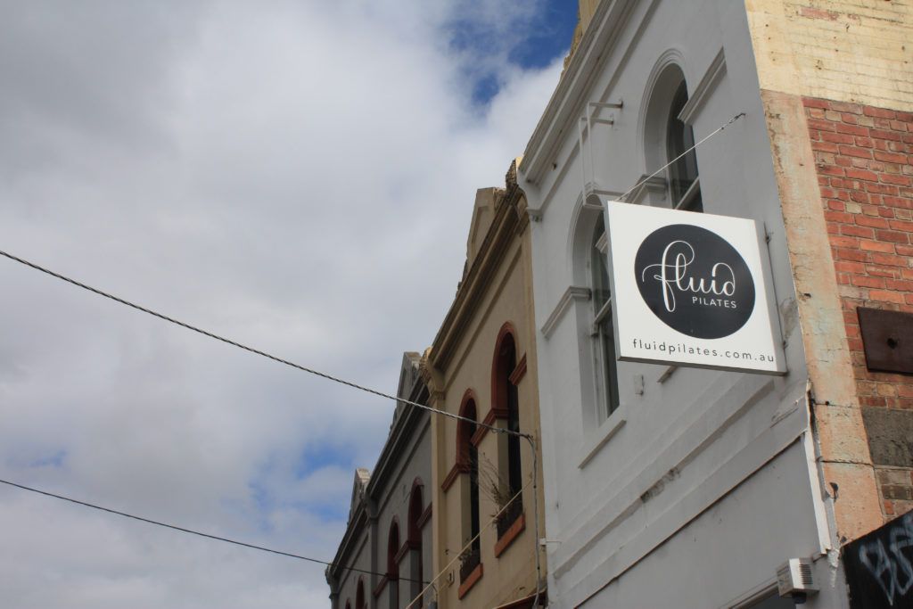 Street corner with a beige brick building and a hanging white shop sign against a cloudy sky