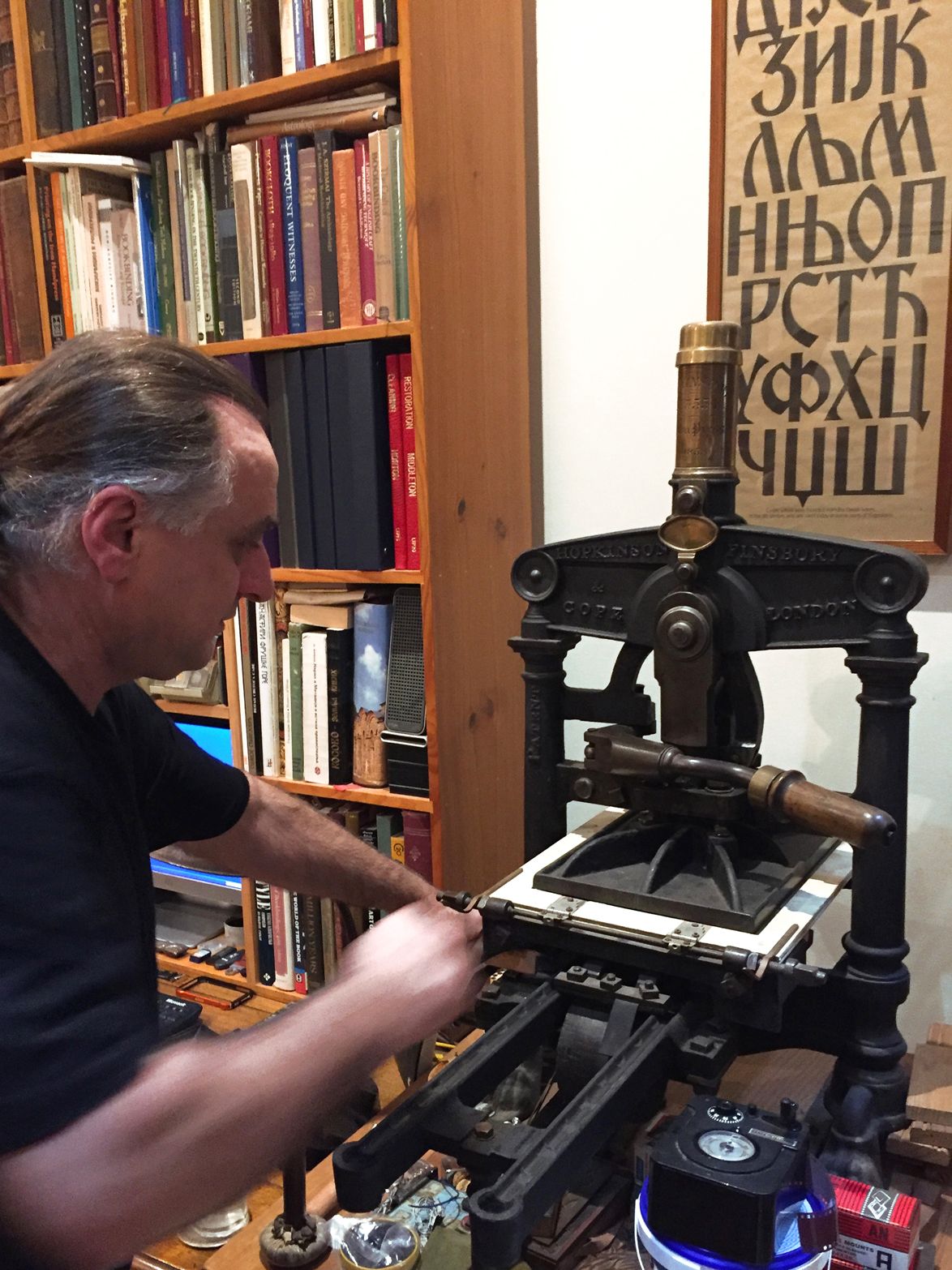 Man operating an antique printing press in a book-lined workshop