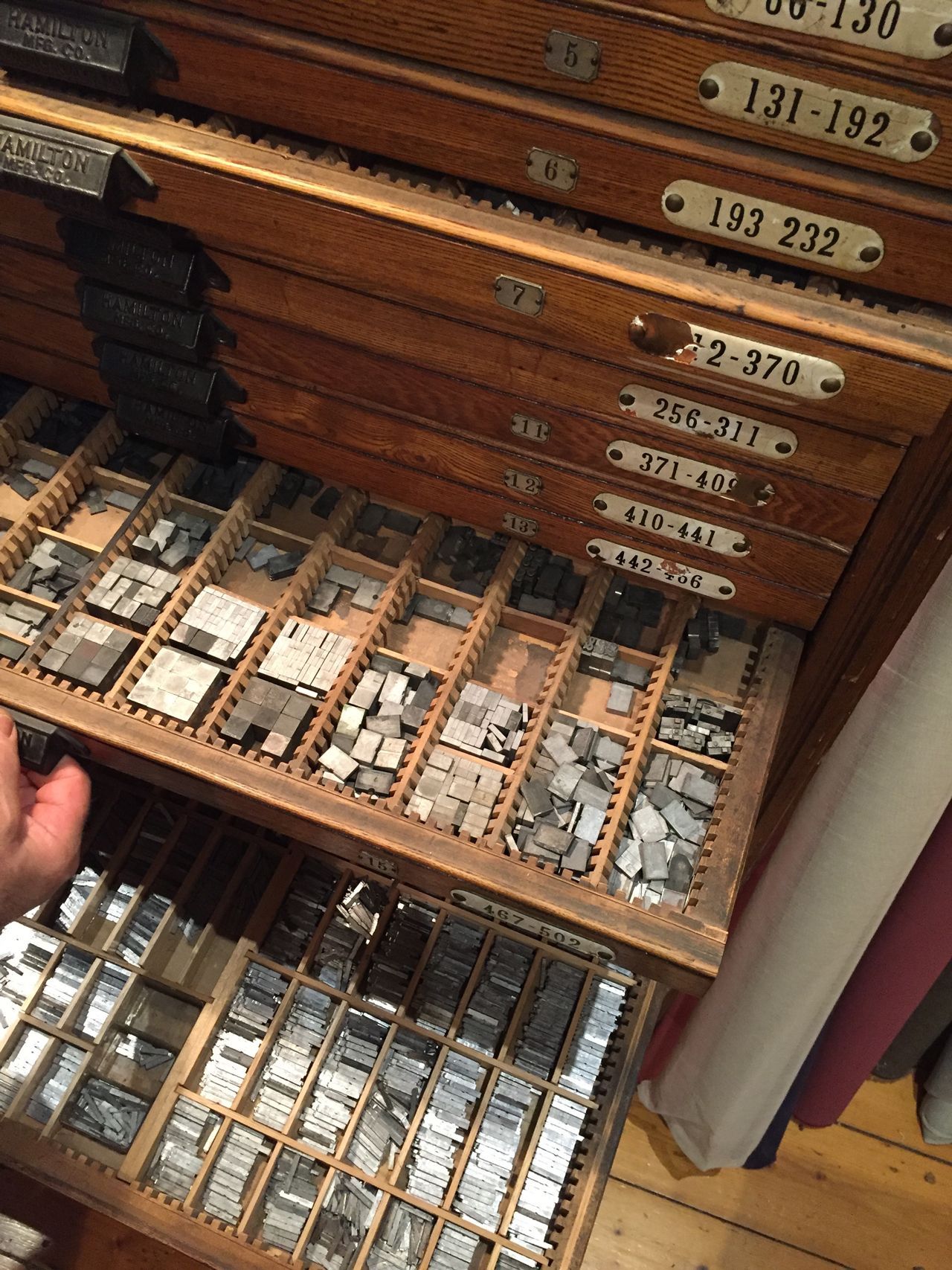 Open drawer of vintage metal type blocks in a wooden cabinet, with a hand reaching in.