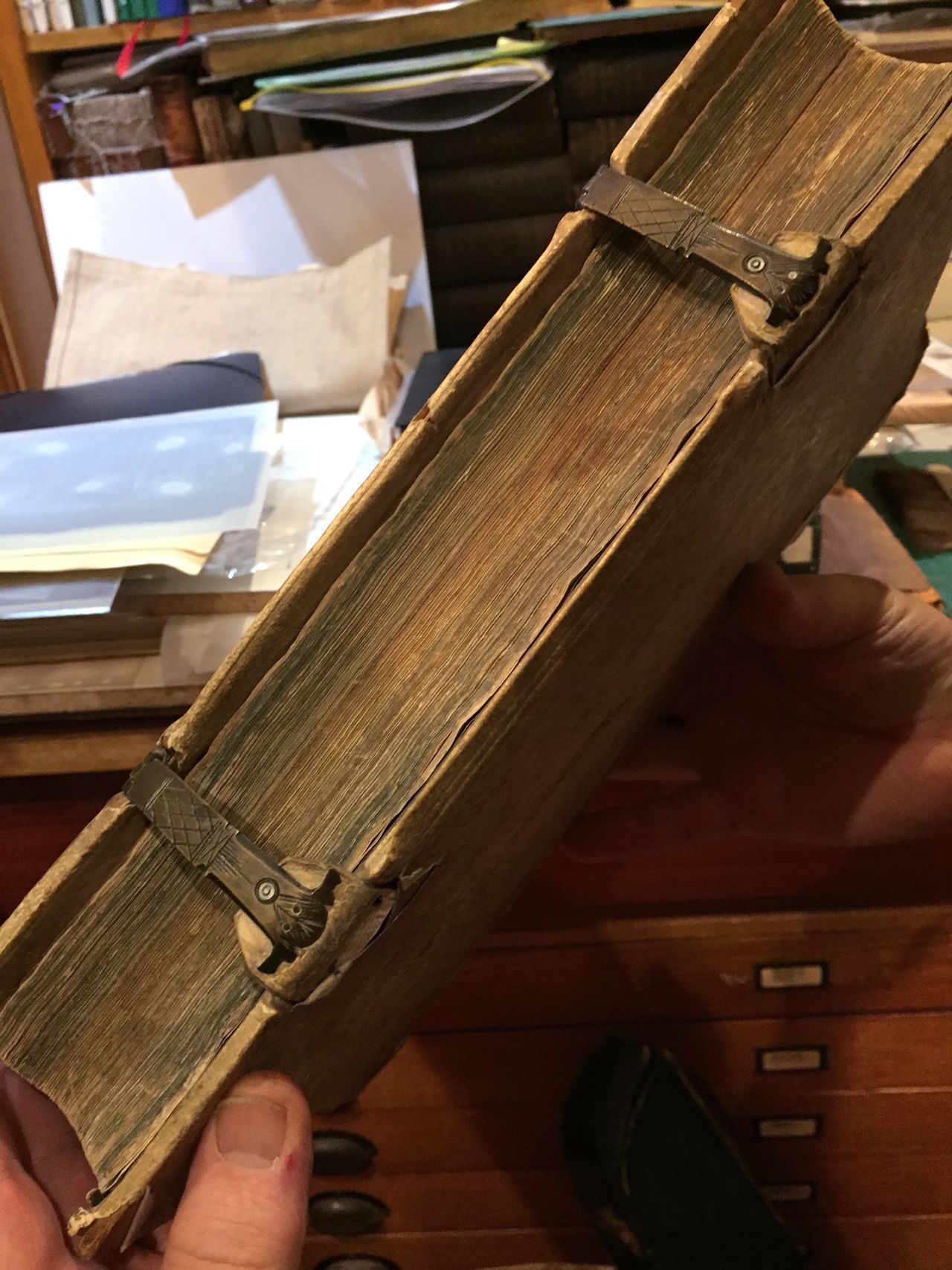 Close-up of a worn, cloth-bound book held in a hand on a cluttered desk