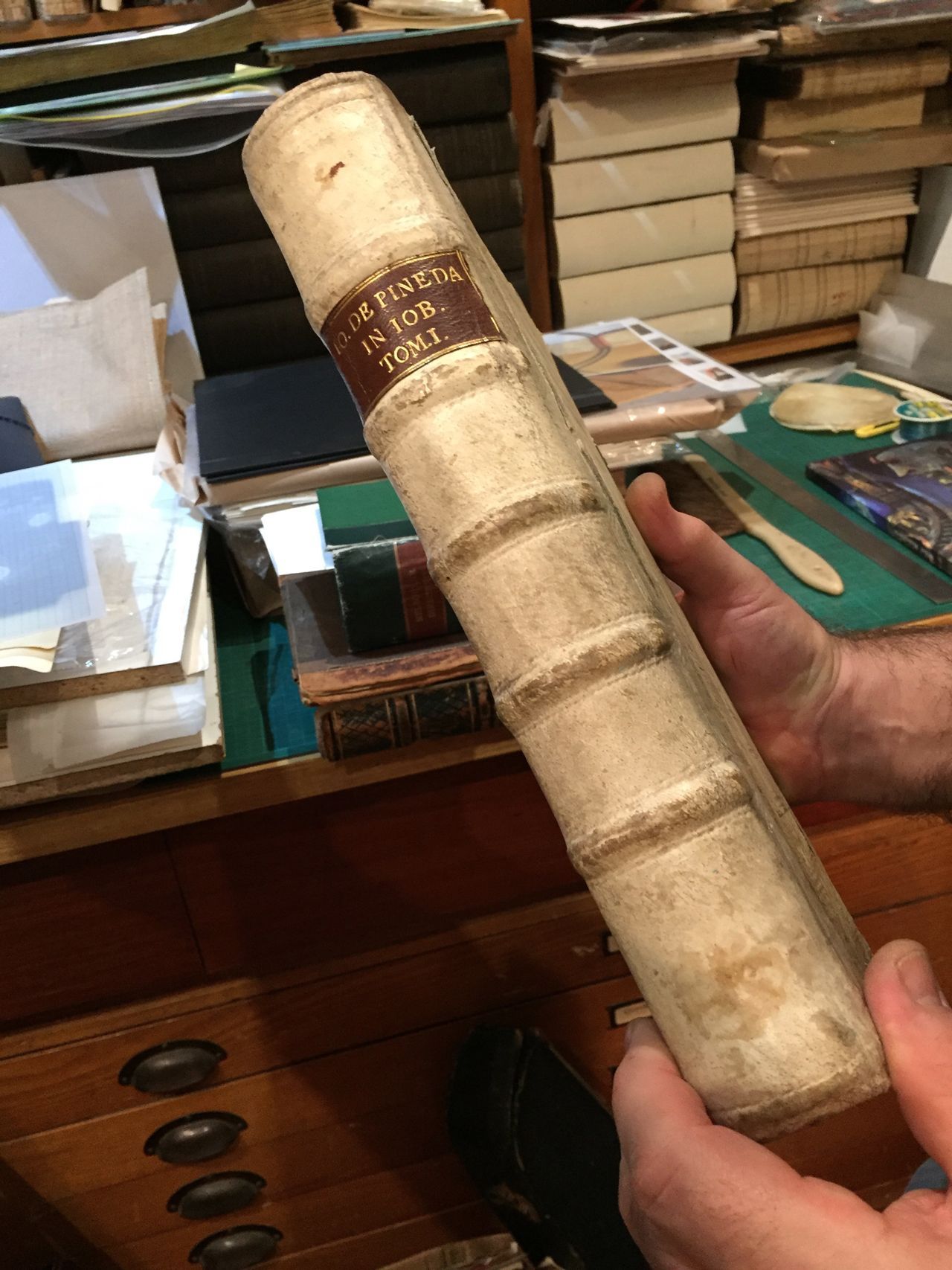 A hand holding a worn beige rolled document or scroll in a cluttered desk setting.