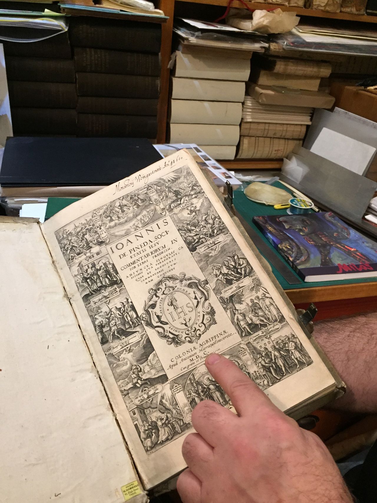 Hand pointing to an open vintage book with black-and-white illustrations on a cluttered table; stacked books behind.