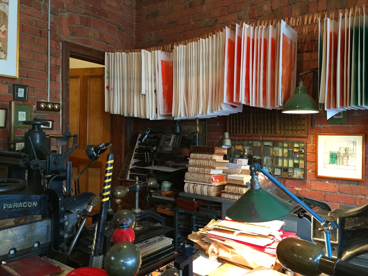 Cluttered workshop with hanging fabric samples, desk lamp, brick walls, and stacked books and supplies