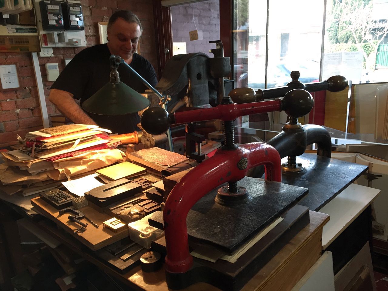 Man operating a press beside stacks of printed cards in a workshop with red equipment