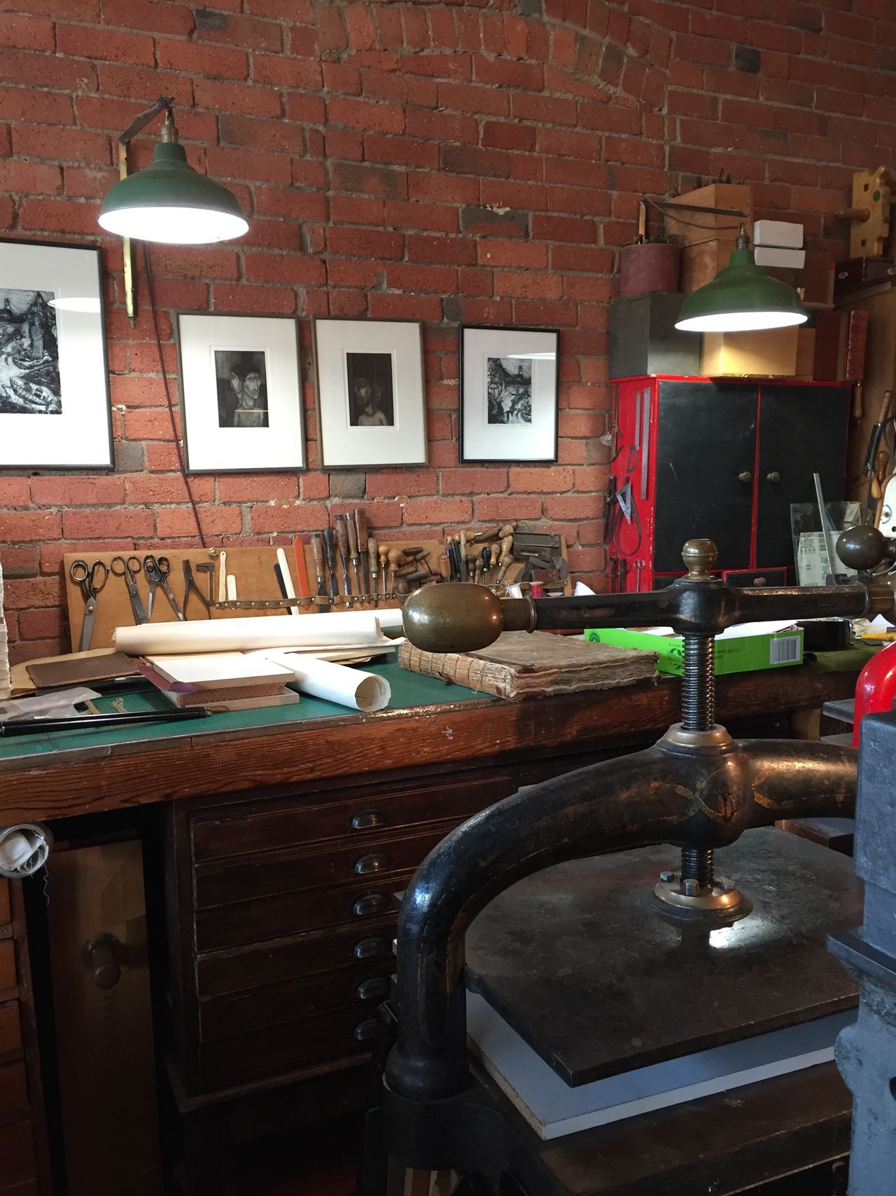 Vintage workshop with brick wall, hanging lamps, framed prints, and a large metal press in the foreground