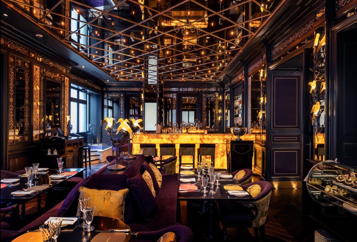 Elegant dimly lit bar and lounge with gold accents, patterned ceiling, and set dining tables