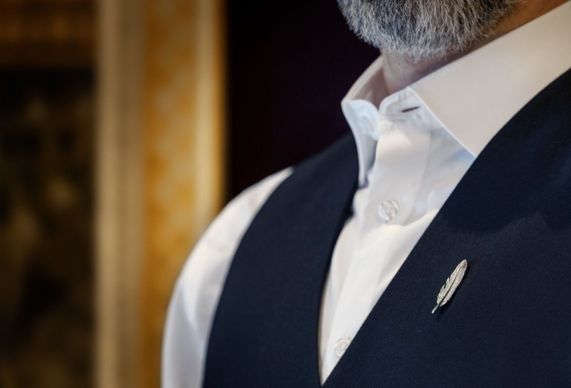Close-up of a man in a white shirt and navy vest with a lapel pin.