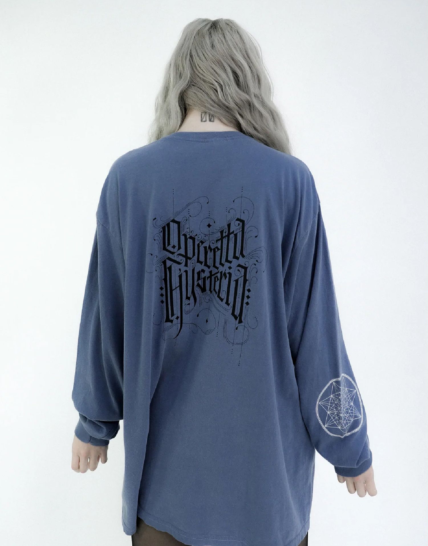 Nina Utashiro seen from behind wearing a blue long-sleeve shirt with black graphic print on a white background