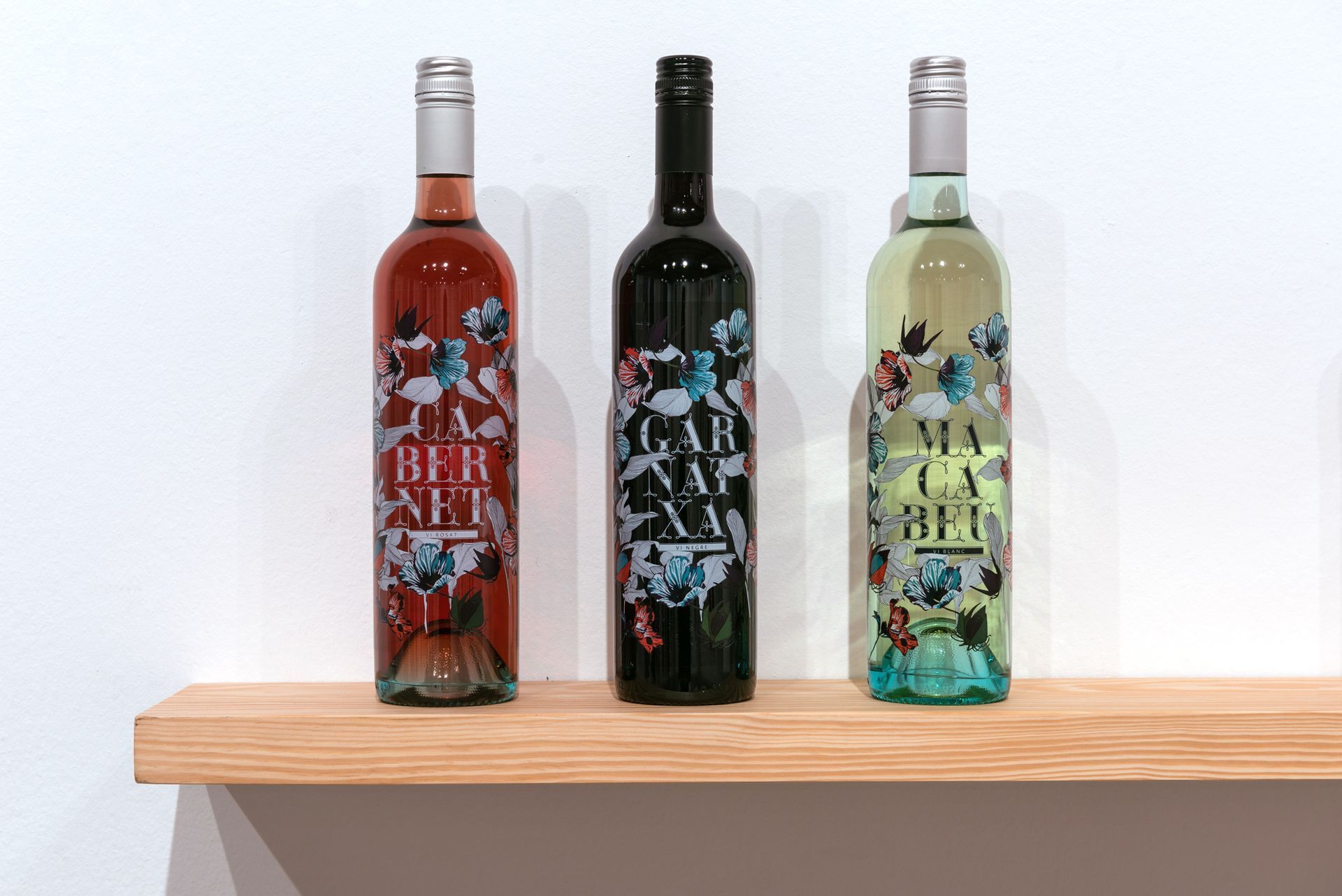Three colorful wine bottles displayed on a wooden shelf against a white wall.