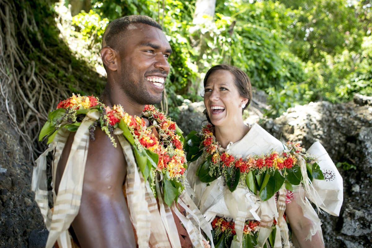 Two smiling people wearing flower leis outdoors in a lush tropical setting