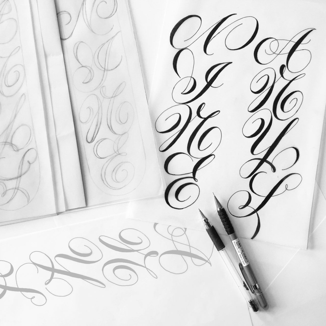Calligraphy practice featuring sheets with intricate flourished letters and two pens resting on a desk.