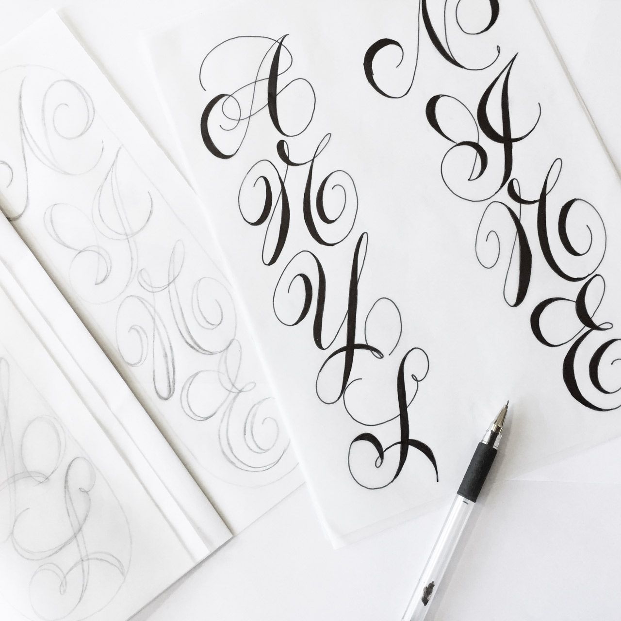 Calligraphic lettering practice on paper with sketches, finished ink letters, and a pen on a white surface.